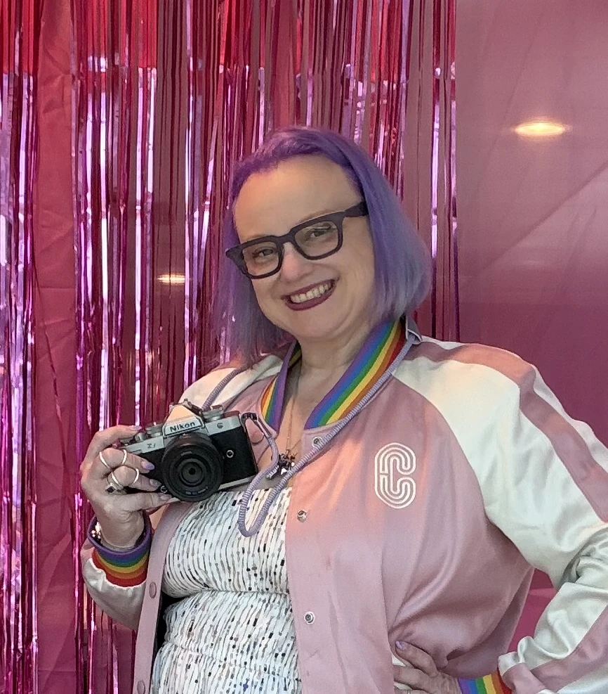 Holding a Nikon camera in a pink jacket, with purple hair and a rainbow camera strap.