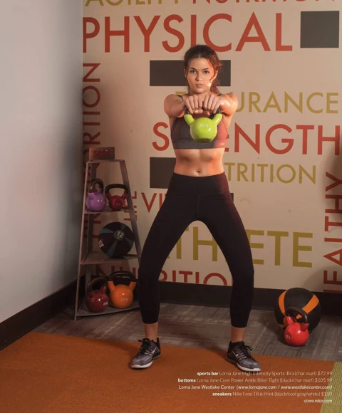 Amanda swinging a green kettlebell at chest height in a charcoal sports bra and black tights, in front of a wall printed with words like PHYSICAL and STRENGTH.