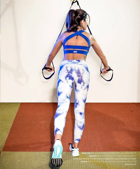 Amanda back-facing on TRX suspension straps, wearing a blue Lorna Jane crossback sports bra and tie-dye Mercury tights against a clean white wall.