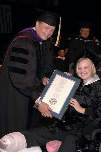 Graduation, May 2005. Jessa in cap and gown receiving a diploma from a faculty member, both feet bandaged in white wraps, seated in a wheelchair.