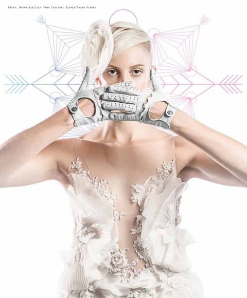 Katie Joy holding white perforated driving gloves over her face, wearing a Luly Yang Couture white lace gown with feather and floral applique, geometric snowflake graphics behind.