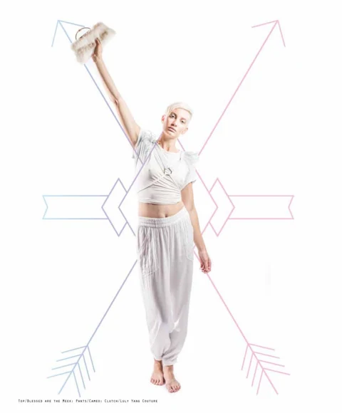 Katie Joy in a draped white Blessed are the Meek crop top, Cameo harem pants, and bare feet, holding a tan fur Luly Yang clutch overhead, large arrow graphics crossing behind her.