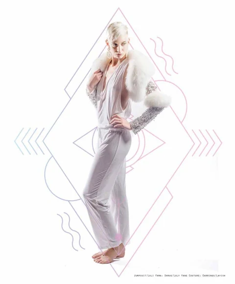 Katie Joy in a Luli Fama white draped jumpsuit and a Luly Yang ostrich-feather and crystal shrug, hand on hip, large diamond graphic outline framing her.