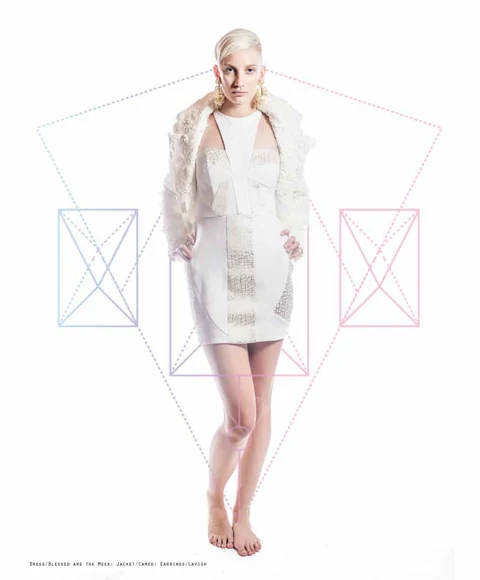 Katie Joy in a white textured Blessed are the Meek mini dress with a Cameo lace-and-bead jacket over her shoulders, geometric kite graphics behind her.