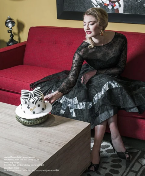 India Gants seated on a red mid-century sofa in a sheer black lace 1950s dress with blue satin bow trim, reaching toward a small white polka-dot fondant cake with a striped bow on the coffee table.