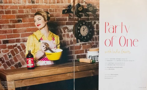Editorial title spread "Party of One with India Gants" — India in a 50s chartreuse button-down dress with a pink polka-dot apron, stirring batter in a bright yellow bowl in a brick-walled kitchen, full credits on the right page.