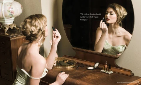 India Gants at a wooden vintage vanity in a light green 1950s slip with embroidered flowers, applying lipstick to her reflection in the round mirror.