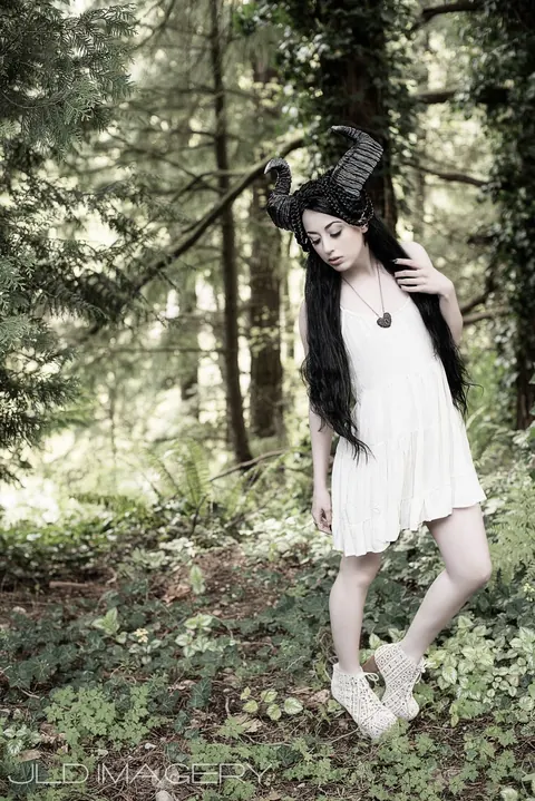 Horned fantasy character in a white dress standing among trees.