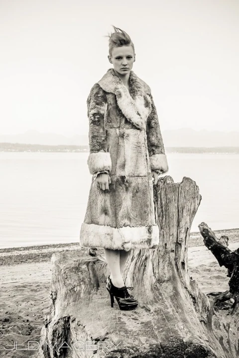 Monochrome fashion portrait in a fur coat beside beach driftwood.