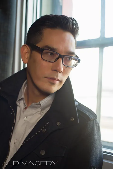 Window-lit portrait of a man wearing glasses and a dark coat.