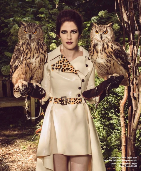 Dawn Mostow in a cream latex trench with leopard print accents, standing in a garden flanked by Lady Bird and Wednesday — two Eurasian Eagle Owls perched on her gloved hands.
