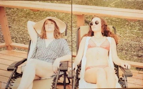 Two women on lakeside chaise lounges, one in a printed romper and floppy sun hat, the other in a coral bikini and crochet vest with round sunglasses.