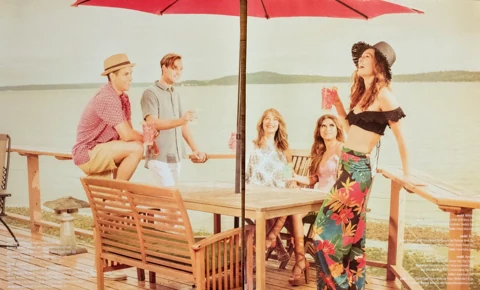 A group of friends gathered around a lakeside table under a red umbrella, one woman in floral wide-leg pants and a black crochet bikini top mid-laugh, holding a drink.