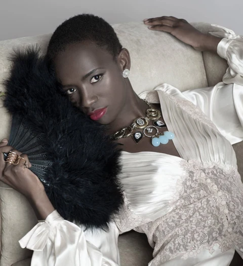 Close-up of Binta Diba reclining in cream silk and lace, holding a black feather fan against her cheek, ornate aquamarine and pearl statement necklace, quiet soft expression.