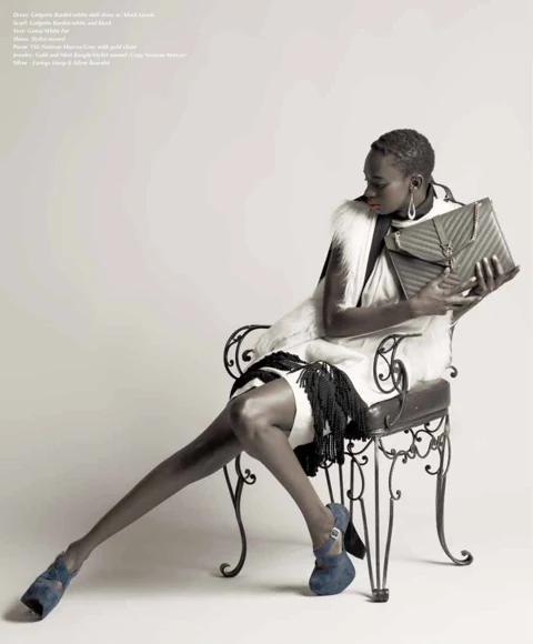 Binta Diba seated on an ornate scrolled iron chair in a Gidgette Bardot white shift dress with black tassel detail, Guess white fur vest, and a quilted YSL clutch held to her face, blue suede platforms on bare floor.