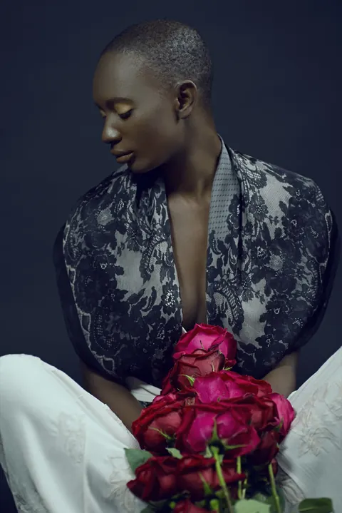 Binta Diba seated in a patterned robe holding a bouquet of red roses.