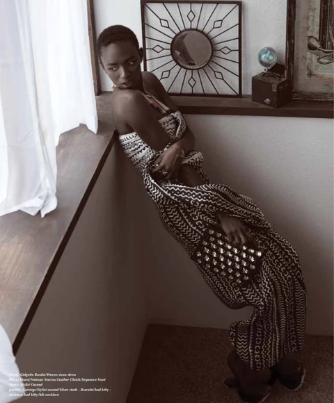 Binta Diba leaning against a half-wall in a black-and-white Gidgette Bardot tribal-print maxi dress with a studded Marni clutch, sunlit window light spilling across the frame.