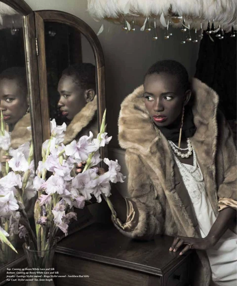 Binta Diba beside a tri-fold vanity mirror in a tan fur coat over an ivory lace gown, layered pearl necklaces, holding a bouquet of pale lavender gladiolas, an ostrich-feather chandelier above.