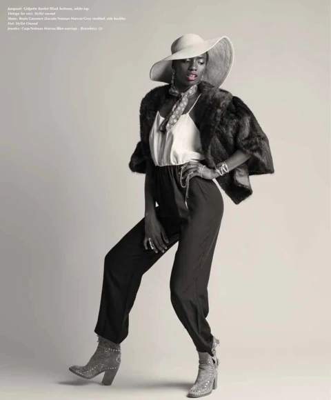 Binta Diba in a wide-brimmed white sun hat, brown faux-fur jacket, ivory satin top, and Gidgette Bardot black jumpsuit with Laurence Dacade studded ankle boots, hand on hip.