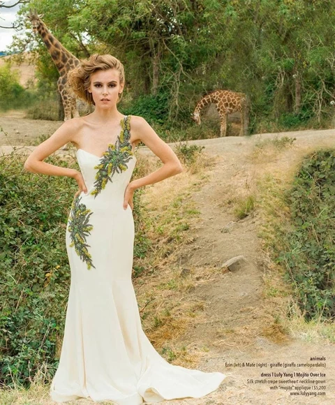 Amberleigh in a one-shoulder ivory Luly Yang gown with green floral applique, standing on a path with Erin and Mate the giraffes browsing trees behind her.